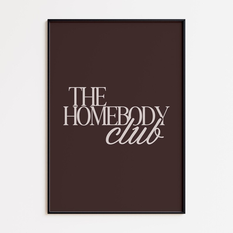 May include: A framed art print with the words "THE HOMEBODY club" in a stylish, light-colored font. The text is centered on a dark brown background, and the frame is black. The print is a decorative piece.