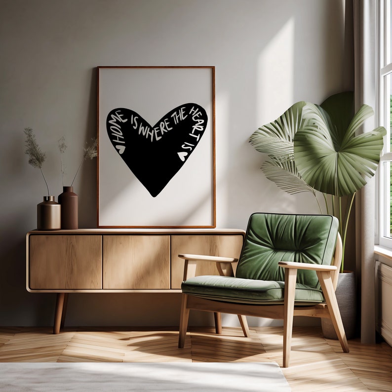 May include: A black and white print with a heart shape and the text "Home is where the heart is". The print is framed in a brown frame and is hanging on a light gray wall. A green armchair and a wooden cabinet are in the background.