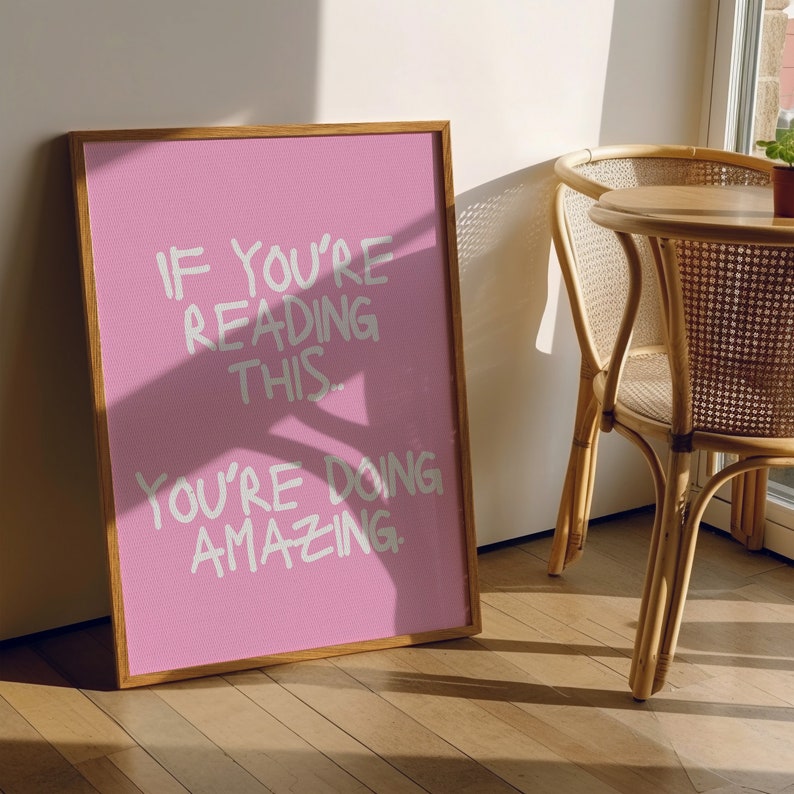 May include: A pink framed poster with the text "If you're reading this, you're doing amazing." The poster is hanging on a wall with a wooden floor and a wicker chair in the background.