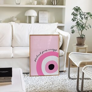May include: Pink and white framed wall art with a pink and white eye design and the text "protect your energy".