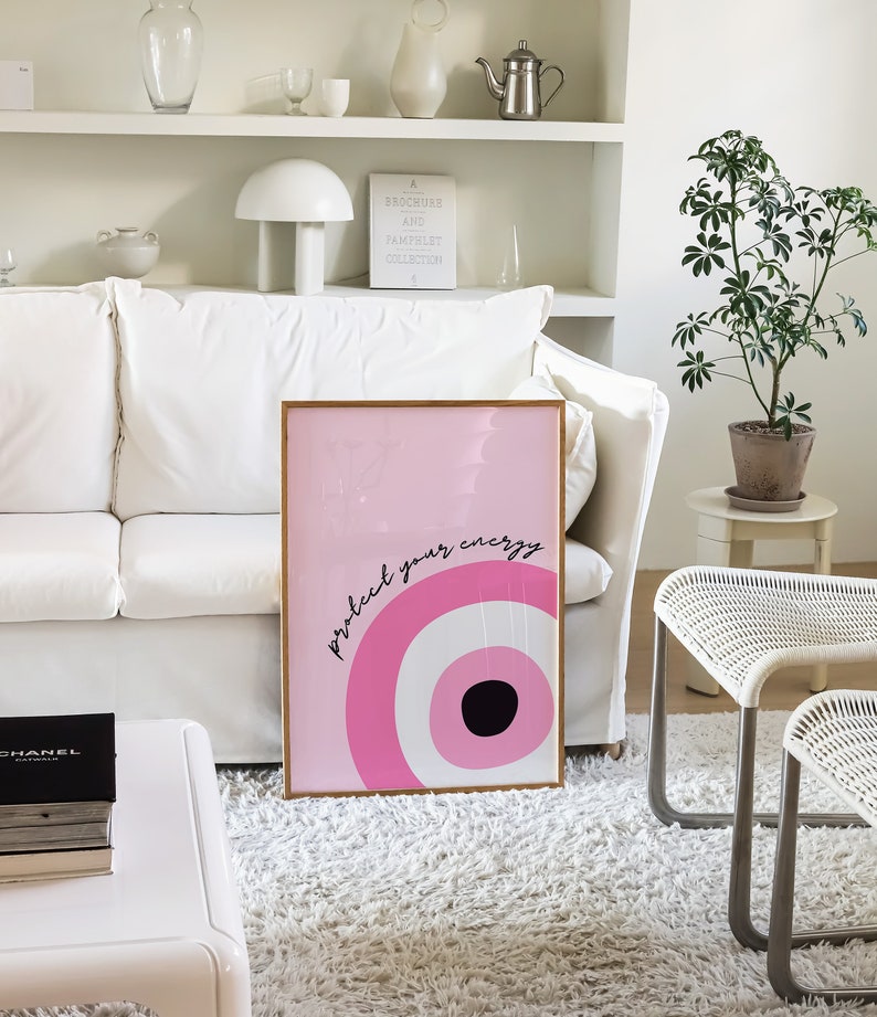 May include: Pink and white framed wall art with a pink and white eye design and the text "protect your energy".