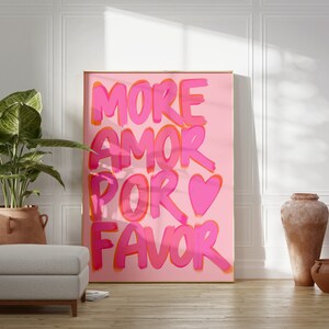 May include: A pink and orange poster with the text "MORE AMOR POR FAVOR" in a bold, playful font. A pink heart is included in the text. The poster is hanging on a white wall in a room with a potted plant and two terracotta pots.
