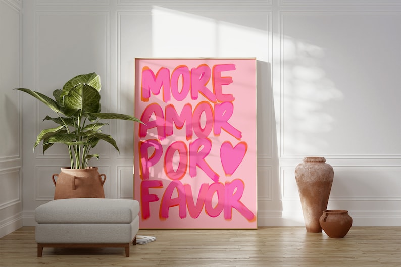 May include: A pink and orange poster with the text "MORE AMOR POR FAVOR" in a bold, playful font. A pink heart is included in the text. The poster is hanging on a white wall in a room with a potted plant and two terracotta pots.