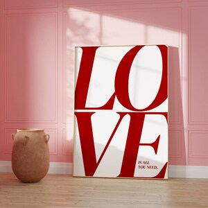 May include: A white canvas print with the word "LOVE" in large, bold, red letters. The text "is all you need." is printed in smaller, black letters below the word "LOVE". The print is framed in a thin, gold frame and is hanging on a pink wall.