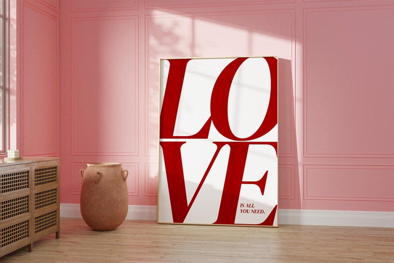 May include: A white canvas print with the word "LOVE" in large, bold, red letters. The text "is all you need." is printed in smaller, black letters below the word "LOVE". The print is framed in a thin, gold frame and is hanging on a pink wall.
