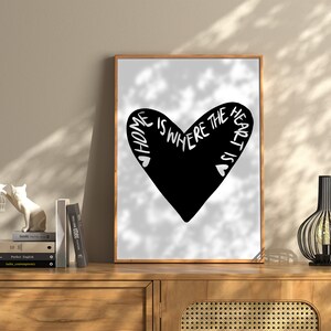 May include: Black and white print of a heart with the text "Home is where the heart is" inside the heart. The print is framed in a light wood frame and is hanging on a wall.