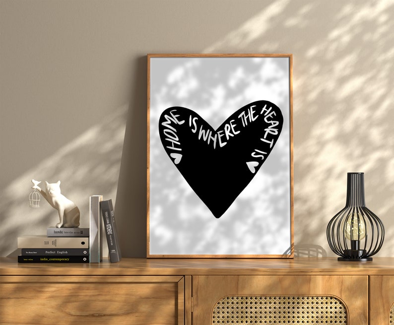 May include: Black and white print of a heart with the text "Home is where the heart is" inside the heart. The print is framed in a light wood frame and is hanging on a wall.