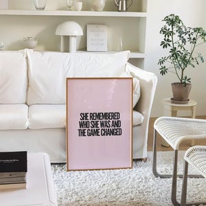 K&ouml;nnte beinhalten: Ein gerahmter rosa Druck mit dem Text "SHE REMEMBERED WHO SHE WAS AND THE GAME CHANGED" in Schwarz. Der Druck lehnt an einem wei&szlig;en Sofa in einem hellen Raum. Eine kleine Topfpflanze und wei&szlig;e M&ouml;bel sind ebenfalls zu sehen.