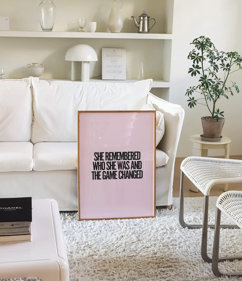 K&ouml;nnte beinhalten: Ein gerahmter rosa Druck mit dem Text "SHE REMEMBERED WHO SHE WAS AND THE GAME CHANGED" in Schwarz. Der Druck lehnt an einem wei&szlig;en Sofa in einem hellen Raum. Eine kleine Topfpflanze und wei&szlig;e M&ouml;bel sind ebenfalls zu sehen.