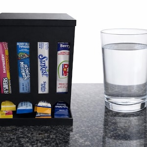 May include: A black box with various drink mix packets, including Fesavors Raspberry Lemonade, Hawaiian Punch, and Sunkist Singles to Go. A clear glass of water sits to the right on a dark gray surface.