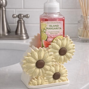 May include: A bottle of Bath & Body Works Island Margarita hand soap sits in a decorative, off-white ceramic holder shaped like sunflowers. The soap bottle is pink with a label showing lime and watermelon slices.