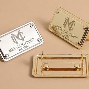 Pode incluir: Três etiquetas metálicas retangulares com cantos arredondados, apresentando o texto gravado "METALLIC CREST est. 1950" e um logótipo de monograma. Uma etiqueta é prateada, duas são douradas, mostrando uma o mecanismo de fixação traseiro.