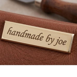 Puede incluir: Primer plano de un artículo de cuero marrón con una etiqueta rectangular dorada. La etiqueta dice "handmade by joe" en una fuente cursiva. El cuero tiene un aspecto texturizado.