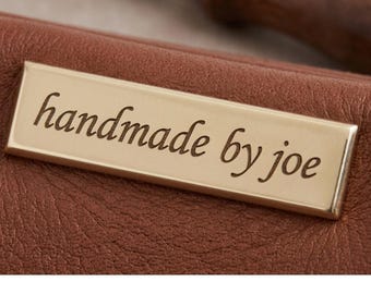 Puede incluir: Un artículo de cuero marrón con una placa metálica rectangular de tono dorado centrada en la superficie. La placa está grabada con el texto negro "handmade by joe" en una fuente cursiva. El cuero tiene un grano visible y una textura suave.