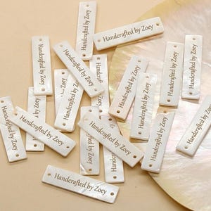 May include: A selection of rectangular, off-white tags, each bearing the inscription "Handcrafted by Zoey" in a serif font. The tags, with two small holes, have a pearlescent finish and are scattered on a light, textured surface.