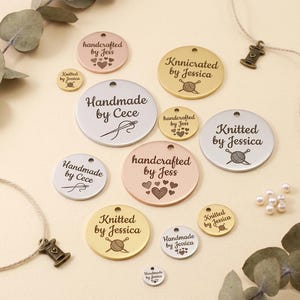 Puede incluir: Una colección de etiquetas redondas de metal en varios tamaños y colores, incluyendo plata, oro y oro rosa. Las etiquetas presentan texto grabado como "Handmade by Cece", "Knitted by Jessica" y "handcrafted by Jess", junto con diseños de corazones y ovillos de lana.