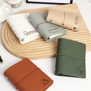 May include: A selection of personalised leather journals in colours such as white, grey, beige, green, and brown. Each journal has a name or initials, secured with an elastic band. The journals are displayed on a wooden chopping board.