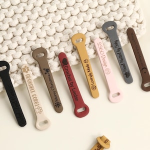 Könnte beinhalten: Eine Sammlung farbenfroher Silikon-Flaschenöffner in verschiedenen Farbtönen, darunter Schwarz, Beige, Braun, Rot, Gelb, Rosa und Grau. Jeder Öffner hat ein einzigartiges Design und Text, wie "Adorable Corgi" und "Merry Christmas".