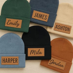 May include: A selection of knitted beanies in shades of green, blue, black, beige, and brown. Each beanie features a rectangular patch with a name: Emily, James, Ashton, Harper, Mila, and Jordan. The beanies are made from a soft, knitted fabric.