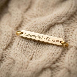 Peut inclure: Une étiquette rectangulaire dorée avec les mots "handmade by Freya" et un symbole de cœur, attachée à un tissu beige en tricot torsadé. L'étiquette a de petits anneaux dorés à chaque extrémité.