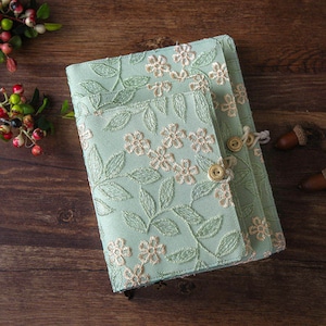 Peut inclure: Une couverture de journal en tissu vert clair avec un motif de broderie florale. La couverture a un rabat avec une fermeture à bouton et un pompon décoratif.