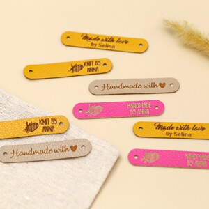 May include: A collection of leather labels in various colors, including yellow, pink, and beige. The labels feature text such as "Made with love by Selina," "Knit by Anna," and "Handmade with." Each label has two holes for sewing.