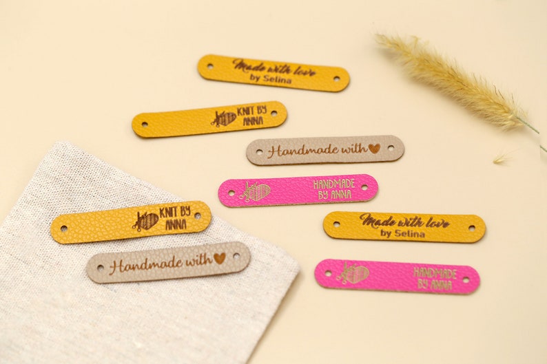 May include: A collection of leather labels in various colors, including yellow, pink, and beige. The labels feature text such as "Made with love by Selina," "Knit by Anna," and "Handmade with." Each label has two holes for sewing.