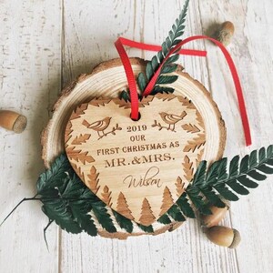 Puede incluir: Un adorno navide&ntilde;o de madera en forma de coraz&oacute;n con el texto "2019 Nuestra primera Navidad como Mr. & Mrs. Wilson" grabado en &eacute;l. El adorno est&aacute; decorado con una cinta roja y un peque&ntilde;o dise&ntilde;o de p&aacute;jaro.