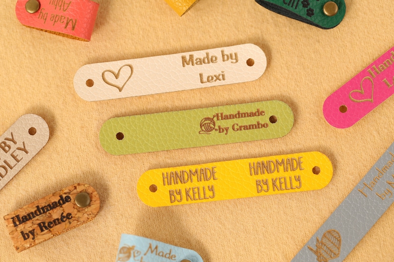 May include: A collection of leather tags in various colors, including pink, green, and yellow. Some tags feature text like "Made by Lexi" and "Handmade by Kelly." Others have heart or yarn ball designs. Each tag has a hole on each end.