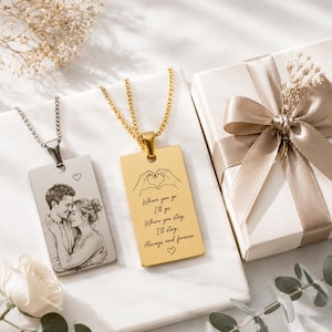 Peut inclure: Deux colliers pendentifs rectangulaires. L'un est argenté avec un portrait de couple et un cœur. L'autre est doré avec le texte "Where you go, I'll go... Always and forever" et un symbole de cœur. Une boîte cadeau avec un ruban est également présente.