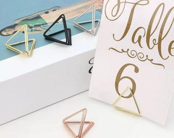 Pode incluir: Uma coleção de porta-cartões geométricos em forma de triângulo em ouro, preto, prata e ouro rosa. Um suporte segura um cartão branco com a palavra "Table" e o número "6" em escrita dourada.