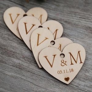 Puede incluir: Un conjunto de etiquetas de madera en forma de corazón. Cada etiqueta está grabada con las letras "V & M" y la fecha "03.11.18". También hay un pequeño corazón grabado. Las etiquetas tienen un agujero para colgar.