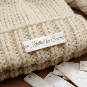 May include: Close-up of a cream-colored knitted hat with a rectangular tag that reads "Knitted by Sue" in a cursive font. The tag is attached to the hat with small holes on each end. Several similar tags are scattered below the hat.