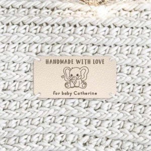Puede incluir: Una etiqueta rectangular de color claro con el texto "HANDMADE WITH LOVE" y "for baby Catherine". La etiqueta presenta una ilustración de un elefante. Está adherida a una tela de punto blanca.