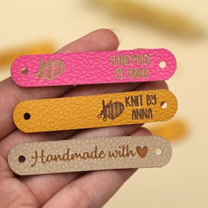 May include: Close-up of three leather tags in pink, yellow, and tan. The pink tag reads "HANDMADE BY ANNA" with a knitting needle and yarn design. The yellow tag says "KNIT BY ANNA". The tan tag says "Handmade with" and a heart symbol.