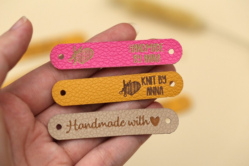 May include: Close-up of three leather tags in pink, yellow, and tan. The pink tag reads "HANDMADE BY ANNA" with a knitting needle and yarn design. The yellow tag says "KNIT BY ANNA". The tan tag says "Handmade with" and a heart symbol.