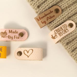 May include: Close-up of several leather tags with text and designs. The tags are in various colors, including pink and tan. Some tags read "Handmade by Alice," "Crocheted by Chey," and "Made by Fia." Other tags feature a heart and a yarn ball design.