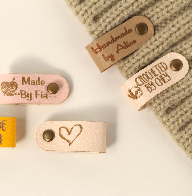 May include: Close-up of several leather tags with text and designs. The tags are in various colors, including pink and tan. Some tags read "Handmade by Alice," "Crocheted by Chey," and "Made by Fia." Other tags feature a heart and a yarn ball design.