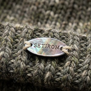 Puede incluir: Primer plano de un gorro de punto en tonos verdes y grises. Una pequeña etiqueta ovalada con la palabra "SETIROM" está adherida al gorro. La etiqueta tiene un acabado nacarado y está asegurada con hilo beige.