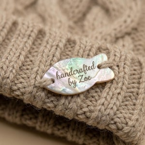 Può includere: Primo piano di un berretto beige a trecce. Un'etichetta madreperlacea è attaccata, con la scritta "handcrafted by Zoe" in corsivo. La trama e il colore neutro del cappello suggeriscono un accessorio fatto a mano e confortevole.
