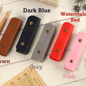 May include: Six leather pencil cases in different colors: brown, dark blue, grey, watermelon red, and pink. The cases are arranged in a row on a white wooden surface.