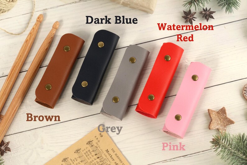 May include: Six leather pencil cases in different colors: brown, dark blue, grey, watermelon red, and pink. The cases are arranged in a row on a white wooden surface.
