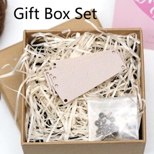 May include: A gift box set with the text "Gift Box Set" includes a light brown box filled with shredded paper, a light pink leather tag, and a small bag of metal hardware. A ball of brown twine is visible.