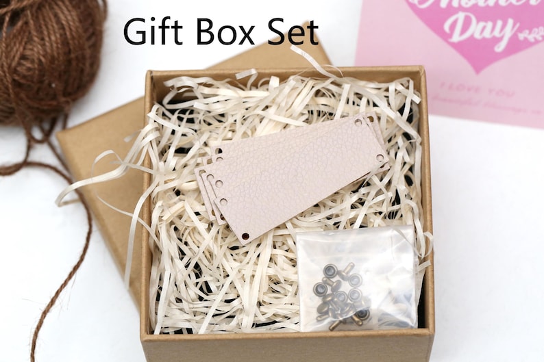 May include: A gift box set with the text "Gift Box Set" includes a light brown box filled with shredded paper, a light pink leather tag, and a small bag of metal hardware. A ball of brown twine is visible.