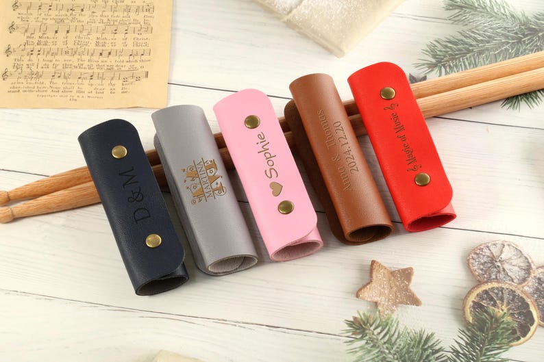 May include: Six personalized leather drum stick holders in various colors, including pink, brown, gray, blue, and red. Each holder has a different inscription, such as "Sophie", "D & M", "2024 12 220", "A Magic of Music", and "Happy Anniversary".