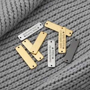 May include: A collection of rectangular metal tags in gold, silver, and gray, some with text. The tags read "made with love Knit by Bella" and "custom knit for Chloe" with heart symbols. The tags are laying on a gray knitted surface.