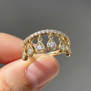 Puede incluir: Un anillo de oro con un diseño único. El anillo presenta una banda curva con pequeños diamantes redondos y colgantes en forma de lágrima, cada uno con un diamante brillante. El anillo es sostenido por una mano contra un fondo borroso.