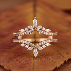 May include: A gold-colored ring with a double band design. The ring features a series of small, clear gemstones and larger marquise-cut stones arranged in a floral or leaf-like pattern. The ring is set against a blurred background.