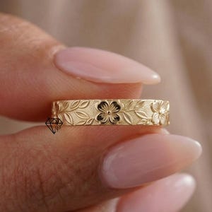 May include: A gold ring with a floral design. The ring has detailed engravings of flowers and leaves. The ring is held between fingers, highlighting the intricate design. The ring's band is approximately 0.5 cm wide, showcasing the craftsmanship.