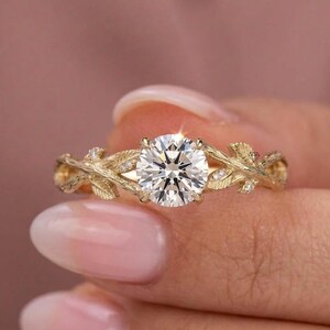 May include: A gold ring featuring a round brilliant-cut center stone held in a four-prong setting. The band is designed with a textured, organic motif resembling intertwined branches and leaves, accented with small, clear, round-cut stones.